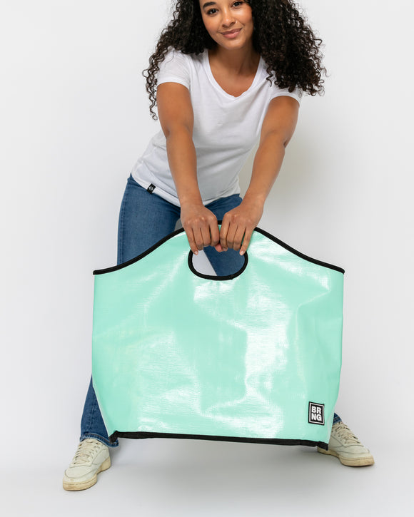 A woman in a white t-shirt and blue jeans holds The Nina Tote by BRNG Bag, a large seafoam green tote with black trim and handles, against a white background—this chic summer bag is perfect for everyday essentials.