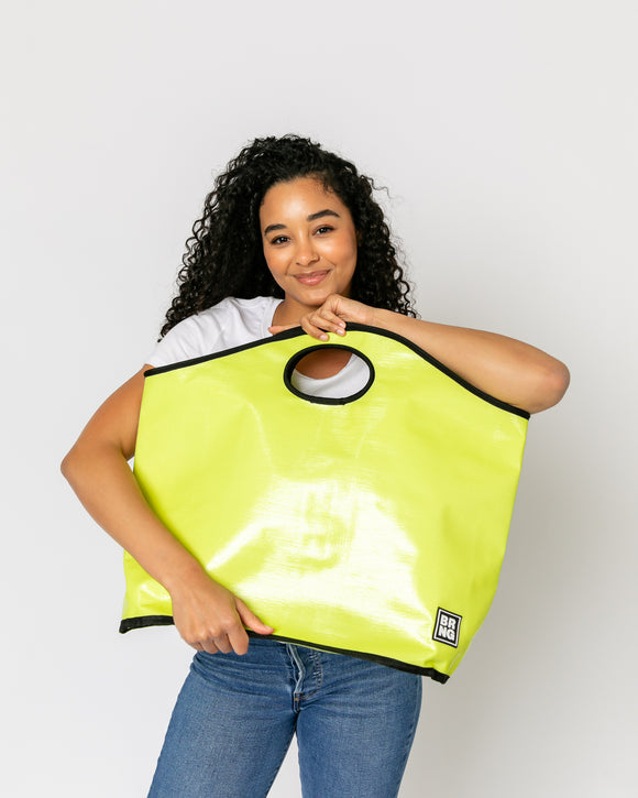 A smiling woman with curly hair and a white shirt holds the BRNG Bag Leni Tote in bright limoncello yellow with black trim and a circular handle, standing against a plain white background and radiating bold style.