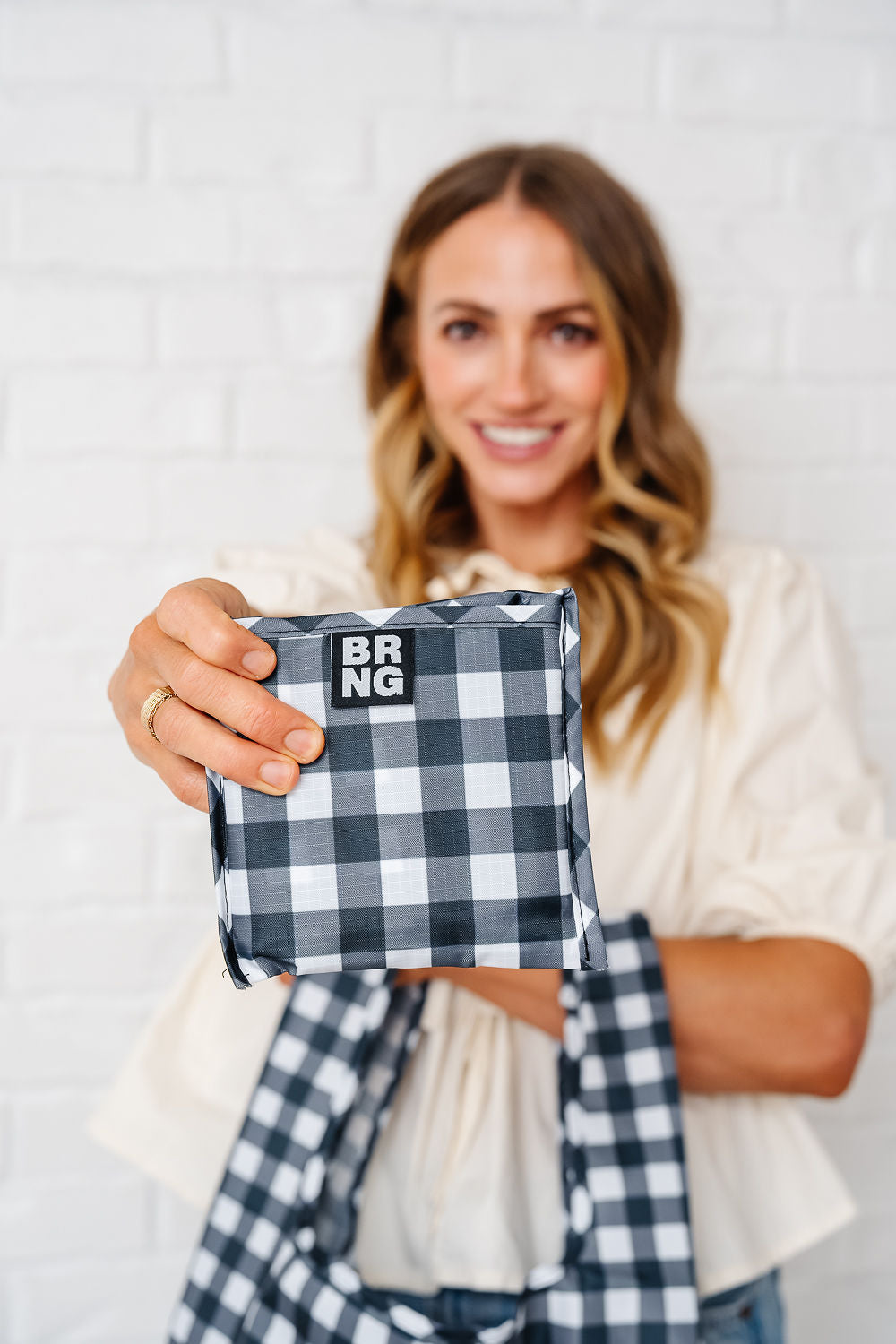 A smiling woman in a cream blouse holds a best-selling black and white checkered Molly Daily shopper by BRNG Bag, featuring the BRNG logo, against a white brick wall background.