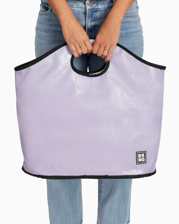 A person in blue jeans holds The Brooke Tote by BRNG Bag—a spacious lavender tote with black trim, handles, and a small BRNG logo at the bottom right corner—covering their waist.
