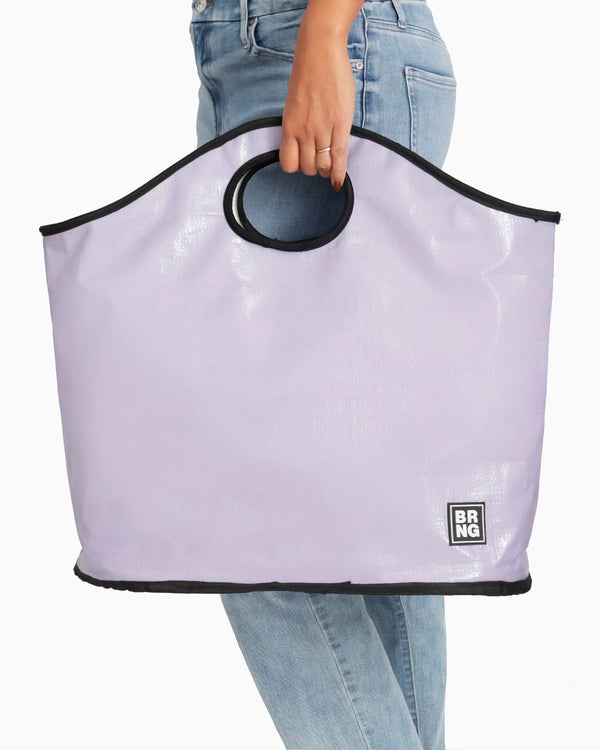 A person in blue jeans carries The Brooke Tote by BRNG Bag—a spacious lavender bag with black trim, circular handles, and a small square BRNG logo near the bottom corner.